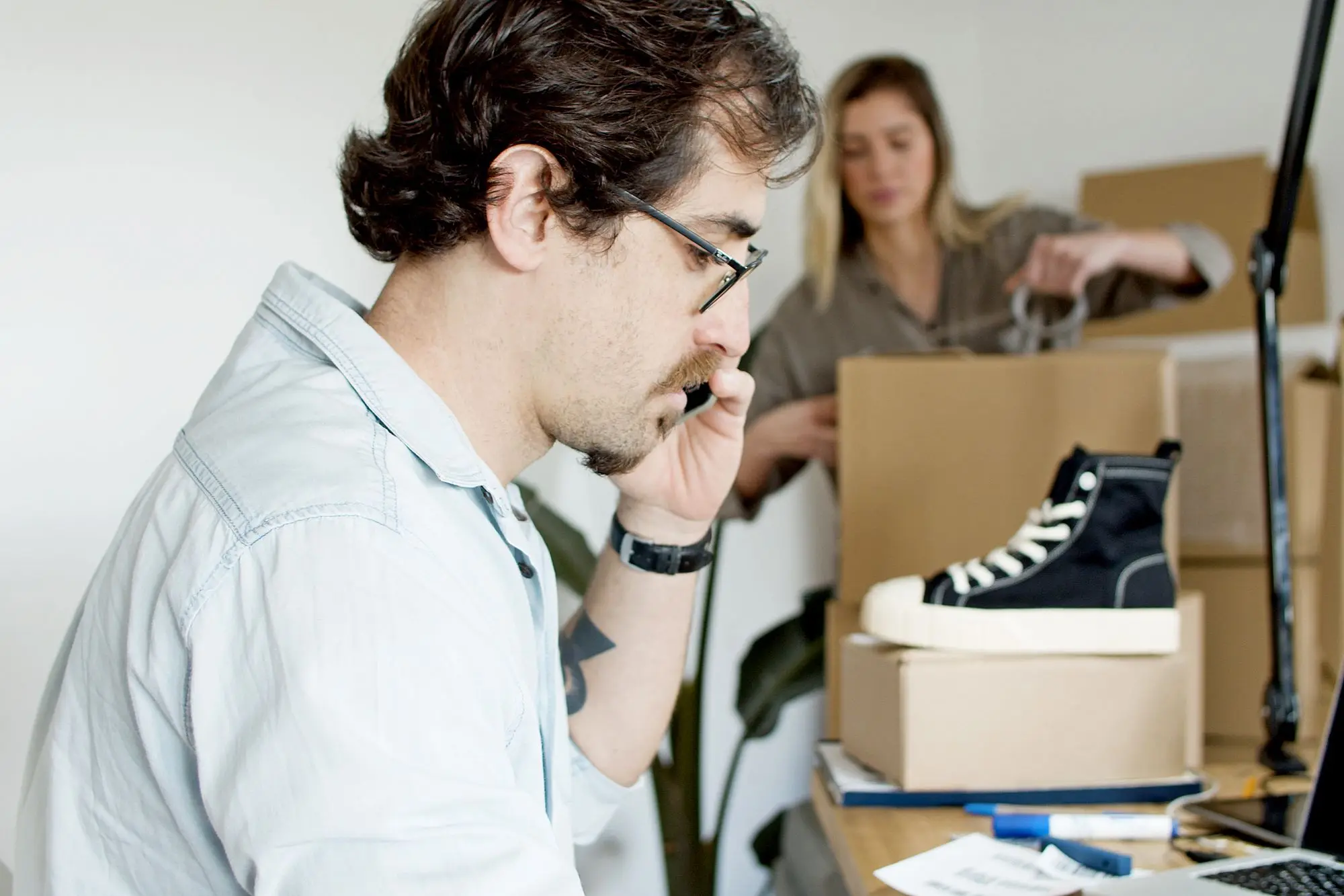 Online store owner on the phone at his desk while a colleague packs sneaker orders into shipping boxes behind him